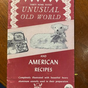 以下が含まれることがあります： 赤い表紙のヴィンテージ料理本。「UNUSUAL OLD WORLD AND AMERICAN RECIPES」と書かれています。表紙には子羊のイラストと食べ物のトレイが描かれています。Nordic Wareのロゴが下にあります。