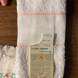 May include: White towel with a pink border and a label that reads "BORDERLINES/FT BF-6662 WHITE/PEACH 100% COTTON Charles Craft Laurinburg, N.C. 28351" and a cross stitch pattern with the text "CROSS STITCH COLLECTIBLES"