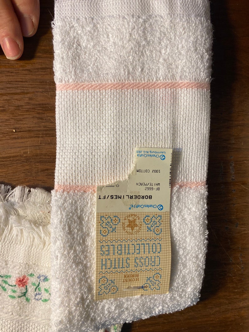 May include: White towel with a pink border and a label that reads "BORDERLINES/FT BF-6662 WHITE/PEACH 100% COTTON Charles Craft Laurinburg, N.C. 28351" and a cross stitch pattern with the text "CROSS STITCH COLLECTIBLES"