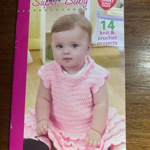 May include: A book titled "Super Baby" featuring a baby in a pink knit and crochet dress. The cover also includes the text "14 knit & crochet projects" and the Red Heart Super Saver logo. The book has a pink spine.