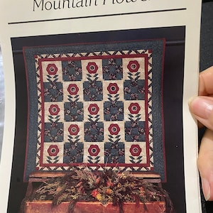 May include: A quilt pattern titled "Mountain Flower" featuring a red, white and blue patchwork design with a floral motif. The quilt is displayed on a wooden shelf with a decorative arrangement of dried flowers and greenery.