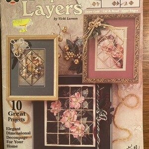 May include: A craft book titled "Paper Layers" by Vicki Larson, featuring 10 great projects for elegant dimensional decoupage. The cover shows three framed images with a gold trim, including a winter scene, a floral arrangement, and a couple in a snowy setting. The book also includes a draw grid, cut and bend instructions, and gold edges.
