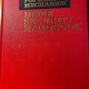 Op de afbeelding: Een rood hardback boek met de titel "HOME SECURITY HANDBOOK" in gouden letters. Het boek is uitgegeven door Popular Mechanics.