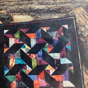 May include: A colorful quilt with a geometric pattern of stars and triangles. The quilt is made of various shades of blue, green, red, and purple fabric. The quilt is set against a dark background.