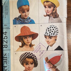 May include: A vintage McCall's sewing pattern for four different hat styles. The pattern is for sizes small and large. The pattern is titled "Hat Wardrobe" and has the number 8254 printed on it. The hats are shown on models with different hair styles and colours. The hats are made of different materials, including straw, felt, and fabric.