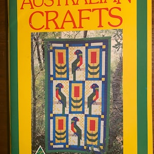 Op de afbeelding: Een boekomslag met een gele achtergrond en de titel "AUSTRALIAN CRAFTS" in grote, vetgedrukte rode letters. De omslag toont een kleurrijk patchwork met een patroon van papegaaien en geometrische vormen. Het patchwork is geplaatst tegen een achtergrond van groene bomen.