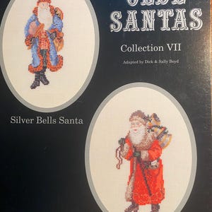 Peut inclure: Un livret de modèles de point de croix de style vintage présentant deux illustrations du Père Noël. Le livret est intitulé "Olde Santas Collection VII" et est adapté par Dick & Sally Boyd. La première illustration montre le Père Noël dans un manteau bleu avec une ceinture rouge et une barbe blanche. La deuxième illustration montre le Père Noël dans un manteau rouge avec une barbe blanche et une bordure en fourrure. Le texte "Silver Bells Santa" est sous la première illustration et "German Series III" est sous la deuxième illustration.