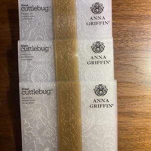 May include: A stack of Cricut Cuttlebug embossing folders with floral and lace patterns. The folders are white and feature the Anna Griffin logo. A tan-colored strip runs down the center of the stack.