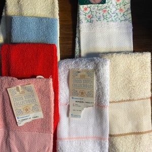 May include: A collection of 10 white, cream, pink, red, and blue towels with fringe edges. The towels have cross stitch designs and labels that say "Cross Stitch Collectibles" and "Borderlines/FT".