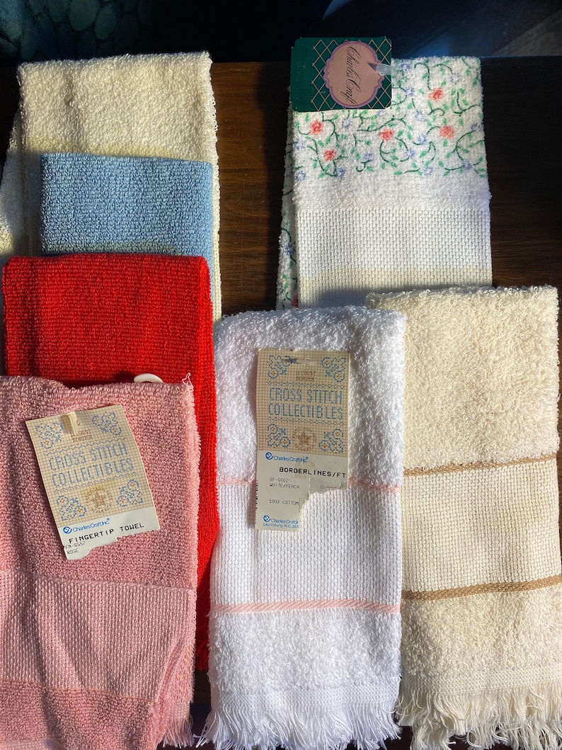 May include: A collection of 10 white, cream, pink, red, and blue towels with fringe edges. The towels have cross stitch designs and labels that say "Cross Stitch Collectibles" and "Borderlines/FT".