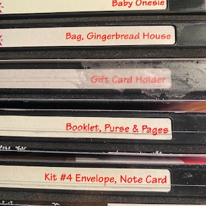 Könnte beinhalten: Fünf schwarze VHS-Kassetten mit weißen Etiketten und rotem Text. Die Etiketten lauten "Baby Onesie", "Bag, Gingerbread House", "Gift Card Holder", "Booklet, Purse & Pages" und "Kit #4 Envelope, Note Card".