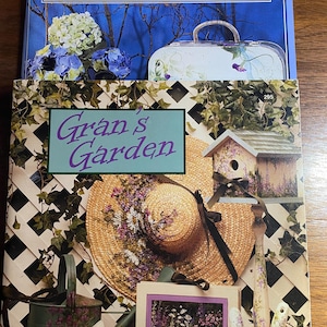 May include: Two books titled "Gran's Memories" and "Gran's Garden" with floral and garden-themed illustrations. The image includes a straw hat, watering cans, and gardening tools, all decorated with flowers. The color palette is soft, featuring blues, purples, and greens.