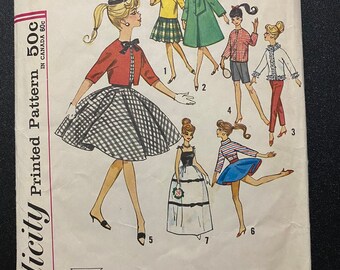 Op de afbeelding: Simplicity naaipatroon voor een pop, nummer 4700. Het patroon bevat instructies voor het maken van zeven verschillende outfits, waaronder een geruite rok, een groene jas, een rood jasje, een witte jurk en een blauwe rok. Het patroon is gedrukt op een witte achtergrond met zwarte tekst.