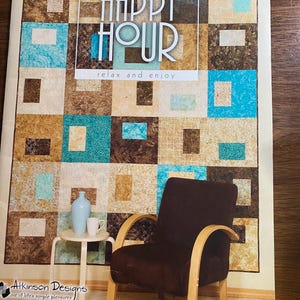 Puede incluir: Un patrón de edredón titulado "Happy Hour" con un diseño de patchwork en tonos marrón, beige y turquesa. La imagen incluye una pequeña mesa con un jarrón azul y una taza blanca, y un sillón marrón con brazos de madera.