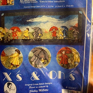 Op de afbeelding: Een kruissteekset met de titel "Slicker Parade" met een kleurrijke illustratie van mensen die onder paraplu's in de regen lopen. De set bevat 14-count navy Aida-stof, een diagram, een naald en voorge sorteerde draden. De afmeting van het afgewerkte ontwerp is 21,5 cm hoog x 57 cm breed.