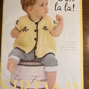 May include: A yellow knitted cardigan with black buttons and leaf embroidery. The cardigan is worn by a baby sitting on a stool. The text "Ooh la la!" is printed on the cover of the book. The book is titled "Tres Fomm Sirdar" and has the number "347 Code B" printed on the top right corner.