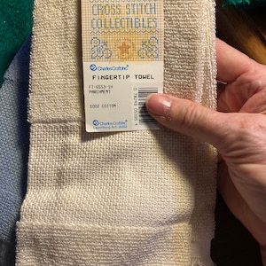 Puede incluir: Una toalla para la punta de los dedos de color pergamino, hecha de 100% algod&oacute;n. La toalla tiene un borde con flecos y una etiqueta que dice "Cross Stitch Collectibles" y "Fingertip Towel". El c&oacute;digo de producto FT-6553-1H tambi&eacute;n es visible.