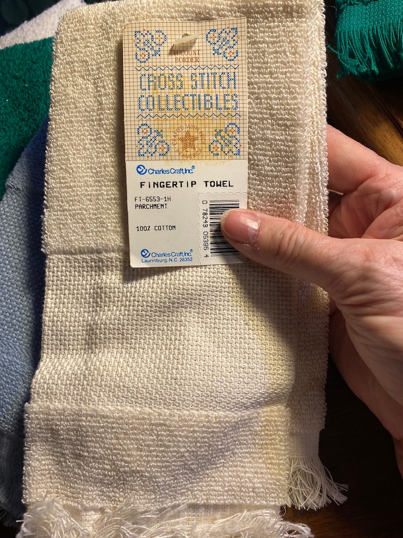 Puede incluir: Una toalla para la punta de los dedos de color pergamino, hecha de 100% algod&oacute;n. La toalla tiene un borde con flecos y una etiqueta que dice "Cross Stitch Collectibles" y "Fingertip Towel". El c&oacute;digo de producto FT-6553-1H tambi&eacute;n es visible.