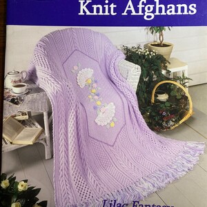Pode incluir: Um cobertor afegão de malha lilás com um design floral, exibido em uma cadeira de vime branca. O cobertor afegão apresenta padrões intrincados e uma borda com franjas. O texto na imagem diz "Award Winning Knit Afghans" e "Lilac Fantasy".