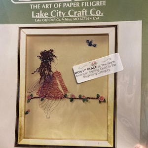 May include: A framed quilling kit featuring a paper filigree design of a fairy. The fairy is depicted in a pink dress and has brown hair. The kit is from Lake City Craft Co. and won 1st place at the North American Quilling Guild.