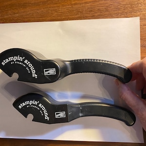 May include: Two black plastic Stampin' Around handle tools for rubber stamps. Each tool features a curved ergonomic grip and a notched head for attaching stamp wheels. The text Stampin' Around by Stampin' Up! is printed in white on the side.