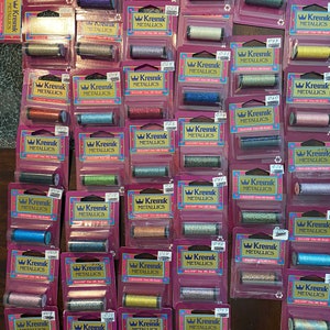 May include: A collection of 24 spools of Kreinik metallic embroidery floss in various colors. The spools are arranged in rows on a wooden surface. Each spool has a label with the Kreinik logo and the words "Kreinik Metallics".