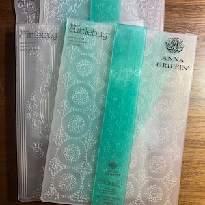 May include: A collection of Cricut Cuttlebug embossing folders and accessories. The folders feature various embossed designs, including floral and geometric patterns. The accessories are a vibrant teal color. The Anna Griffin brand is visible on some of the folders.