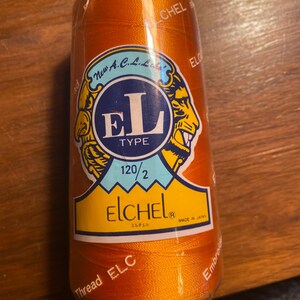 Op de afbeelding: Een spoel oranje borduurgaren met het label "ELCHEL" en de tekst "New A.C.L. Laks" en "EL TYPE 120/2" gedrukt op het label. De spoel is gemaakt in Japan.