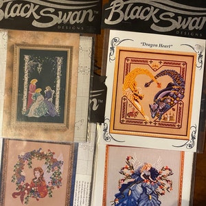 May include: Two cross-stitch patterns from Black Swan Designs. The first pattern features two women in long dresses standing in a garden. The second pattern features a dragon heart with a yellow and purple dragon. The third pattern features a fairy with blue wings and a blue dress.