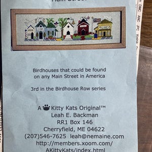 May include: A cross-stitch pattern titled Birdhouse Row #3 Main Street featuring a row of colorful small buildings. The text includes Birdhouses that could be found on any Main Street in America, 3rd in the Birdhouse Row series, and A Kitty Kats Original by Leah E. Backman with contact details.
