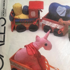 May include: A toy train set with a red engine, a blue boxcar, and a pink unicorn boxcar. The engine has a yellow boxcar attached to it. The train is made of fabric and has a circus train design.