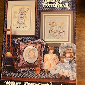 Pode incluir: Uma capa de livro de padrões de ponto cruz intitulada "Dolls of Yesteryear" com o subtítulo "Stoney Creek Collection." A capa apresenta uma imagem de três bonecas, duas sentadas e uma em pé, e um padrão de ponto cruz de uma menina com um cordeiro e uma menina em uma carruagem. O livro é o número 69 e custa 5,00 €.