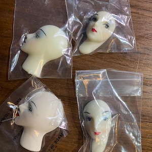 Puede incluir: Cuatro cabezas de muñeca vintage, cada una en una bolsa de plástico transparente. Las cabezas de muñeca son de color blanquecino con rasgos pintados, incluyendo cejas y pestañas negras y labios rojos. Las cabezas están en varias orientaciones.