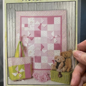 May include: A pink and white patchwork quilt with a green and pink tote bag, two teddy bears, and a wicker basket. The quilt is made with a variety of fabrics, including floral prints and gingham. The tote bag has a green and white windmill design. The teddy bears are sitting on top of the basket. The image includes the text "#1146 Charming Baby".