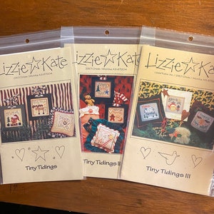 Może przedstawiać: Trzy brązowe papierowe torby z białym tekstem i wzorami gwiazd. Torby są oznaczone jako "Tiny Tidings", "Tiny Tidings II" i "Tiny Tidings III". Każda torba zawiera inny wzór haftu krzyżykowego o tematyce świątecznej.