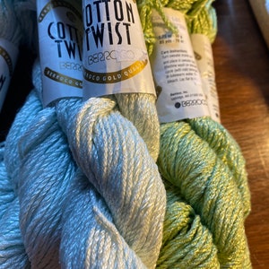 May include: Close-up of several skeins of Berroco Cotton Twist yarn in shades of light blue and green. The yarn is labeled with the brand name and product details. The skeins are tightly wound and ready for crafting.