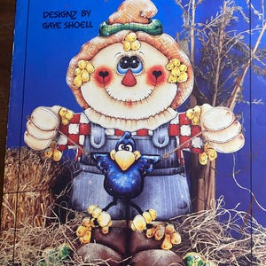 Puede incluir: Ilustración colorida de un espantapájaros llamado "Mr. Quackenbush" con un pájaro azul. El espantapájaros tiene un sombrero de paja, una camisa a cuadros rojos y blancos y un peto azul. La obra es de Gaye Shoell.