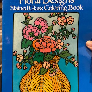 May include: A blue book titled "Floral Designs Stained Glass Coloring Book" by Ed Sibbett, Jr. The cover features a floral design in a woven basket, with pink and orange flowers and green leaves. The Dover Coloring Book logo is in the upper left corner.
