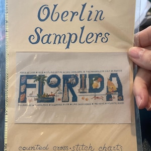 May include: A cross-stitch chart from Oberlin Samplers featuring the word "FLORIDA" in blue, with various Florida landmarks and locations depicted within the letters. The chart is designed by M.M. Waldock.