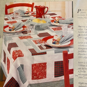 Könnte beinhalten: Eine weiße Tischdecke mit rotem und schwarzem Karomuster. Die Tischdecke ist mit roten und weißen Quadraten mit einem festlichen Design verziert. Die Tischdecke ist für ein festliches Abendessen mit roten und weißen Servietten, Gläsern und einer roten Kanne gedeckt.