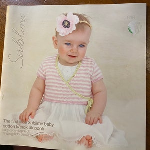 Puede incluir: Una portada de libro con un bebé que lleva un suéter de punto a rayas rosa y blanco y un vestido blanco. El título del libro es "The first little Sublime baby cotton kapok dk book" y el subtítulo es "14 designs for babies from 0 to 3 years".