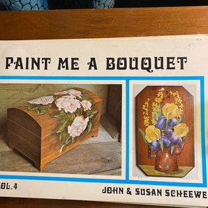 May include: A book titled "Paint Me A Bouquet" Vol. 4 by John & Susan Scheewe. The book features two images of painted wooden crafts. The first image shows a wooden box with a painted bouquet of pink roses. The second image shows a wooden plaque with a painted bouquet of yellow and purple flowers in a brown teapot.