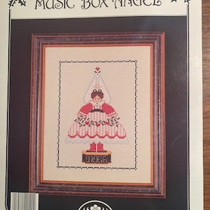 May include: A cross stitch pattern for a music box angel. The pattern features a red and white dress with a holly border. The angel is standing on a music box with the word "music" written on it. The pattern is from Book 75, HS120, Astor Place.