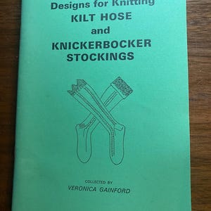 Könnte beinhalten: Ein grünes Heft mit dem Titel "Designs for Knitting Kilt Hose and Knickerbocker Stockings" und einer Zeichnung von gekreuzten Strümpfen. Das Heft wurde von Veronica Gainford zusammengestellt.