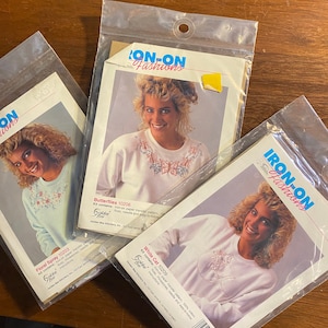 May include: Three iron-on transfer pattern kits for clothing. Each kit includes a white t-shirt with an embroidered design, iron-on paper transfer pattern, rhinestones, needle, and easy-to-follow instructions. The designs are Butterflies, Floral Spray, and White Cat.