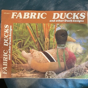 以下が含まれることがあります： 「FABRIC DUCKS and other Duck designs」というタイトルの書籍で、表紙には布製の鴨が描かれています。鴨は、茶色、緑色、赤色で、ペイズリー柄になっています。