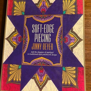May include: Book cover with a purple starburst design and the title "Soft-Edge Piecing" by Jinny Beyer. The subtitle reads "Add the elegance of appliqué to traditional-style patchwork design."