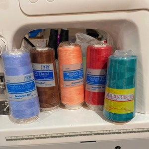 Może przedstawiać: Pięć szpulek nici do overlocka w różnych kolorach, w tym niebieski, brązowy, brzoskwiniowy, czerwony i turkusowy. Każda szpulka jest oznaczona "Overlock Thread" i "Beachwood Ltd."