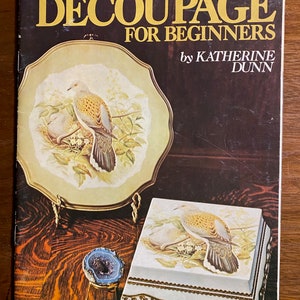 May include: A book titled "Decoupage for Beginners" by Katherine Dunn, featuring a cover image of a decorative plate and a small box with a dove design. The book is priced at one dollar and twenty-five cents.