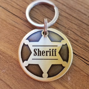 May include: A circular brass-toned metal keychain tag featuring a six-pointed star cutout design. The word Sheriff is engraved in black lettering across the center of the star. The tag is attached to a silver-colored metal split ring for keys.